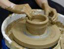Ruce potřené mokrým jílem tvarují keramickou nádobu na hrnčířském kruhu. Hlína se otáčí na kole a okraje nádoby jsou jemně upravovány. Kolem kruhu je vidět rozstříkaný jíl a pracovní nepořádek.